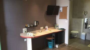 Una pequeña kitchenette con cafetera, una estantería con libros, un cubo de basura y un inodoro visible a través de una puerta abierta.