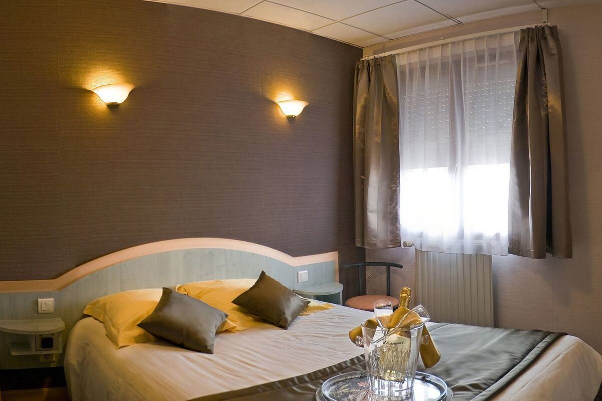A hotel room with a bed, two wall-mounted lights, a window with curtains, a bedside table, and a tray with a bottle and glasses.