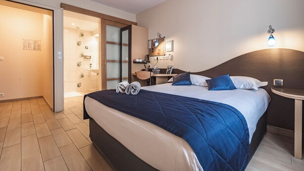 A hotel room with a large bed, a desk, a chair, a small table, a wall-mounted lamp, and a door leading to a bathroom. - Logis Hôtel Au Tambour (Reims)