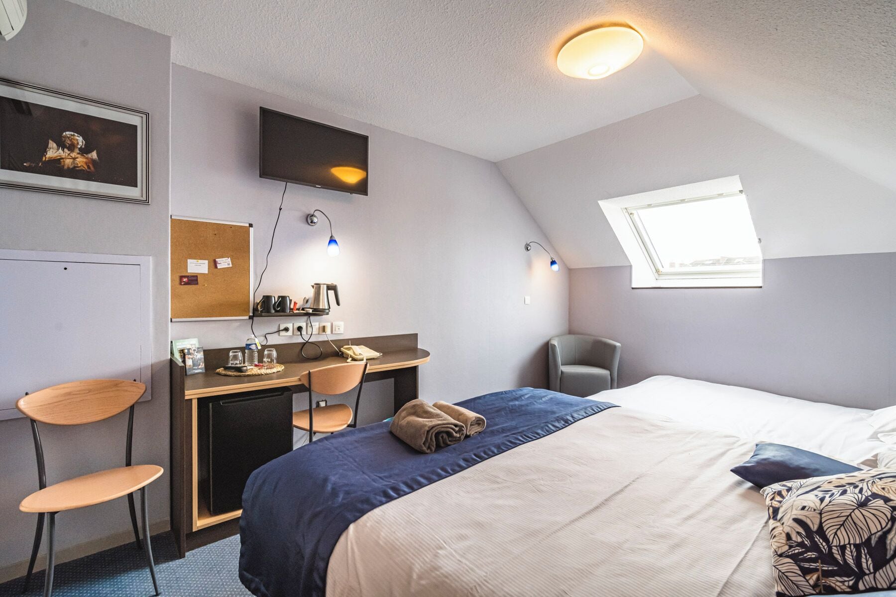A bedroom with a bed, desk, chairs, a television, a window, and a skylight.