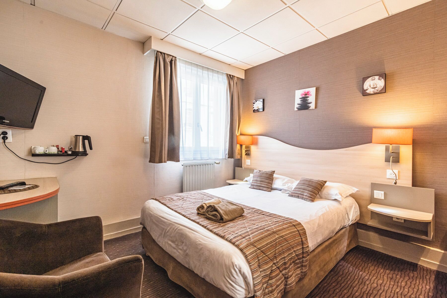 A hotel room with a bed, bedside tables, a desk with a coffee maker, a TV, and a window with curtains.