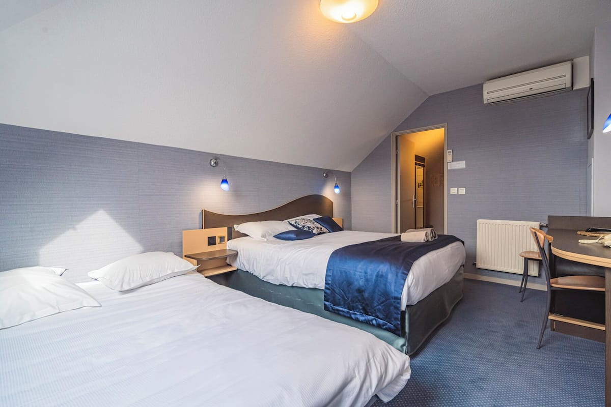 A hotel room with two beds, a desk, and a chair. There is a radiator on the wall and an air conditioning unit above the door.
