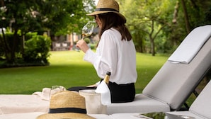 A person seated on a grey lounge chair outdoors, holding a glass of white wine.