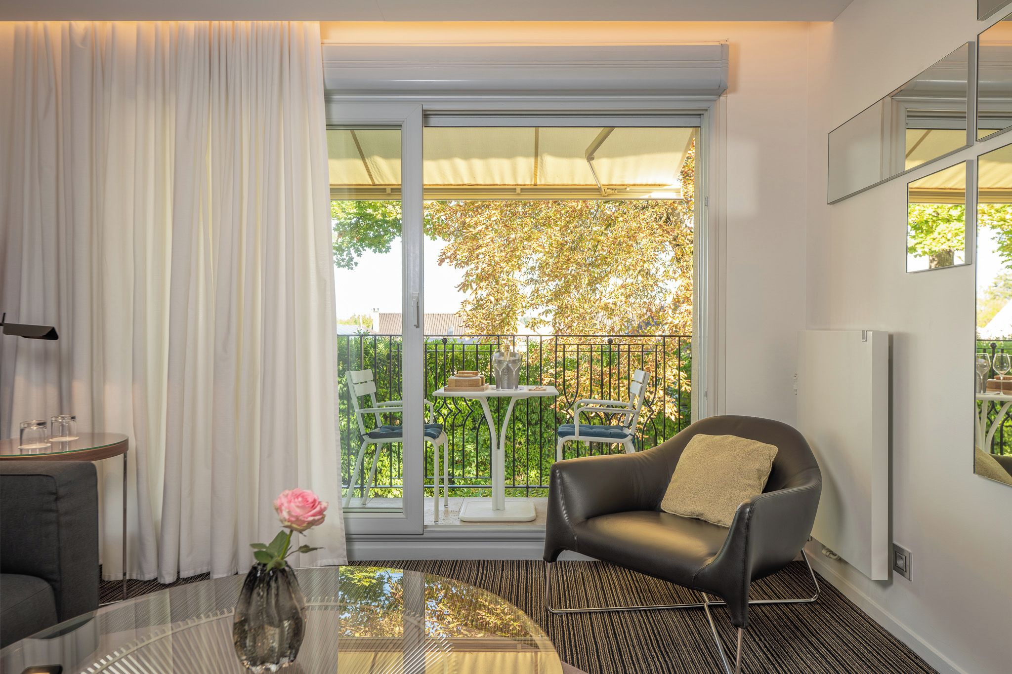 A modern living room with a glass table, a leather armchair, and a view of a balcony with outdoor furniture.