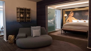 A modern hotel room with a large bed, a sofa, a small shelf, and a view of the city.