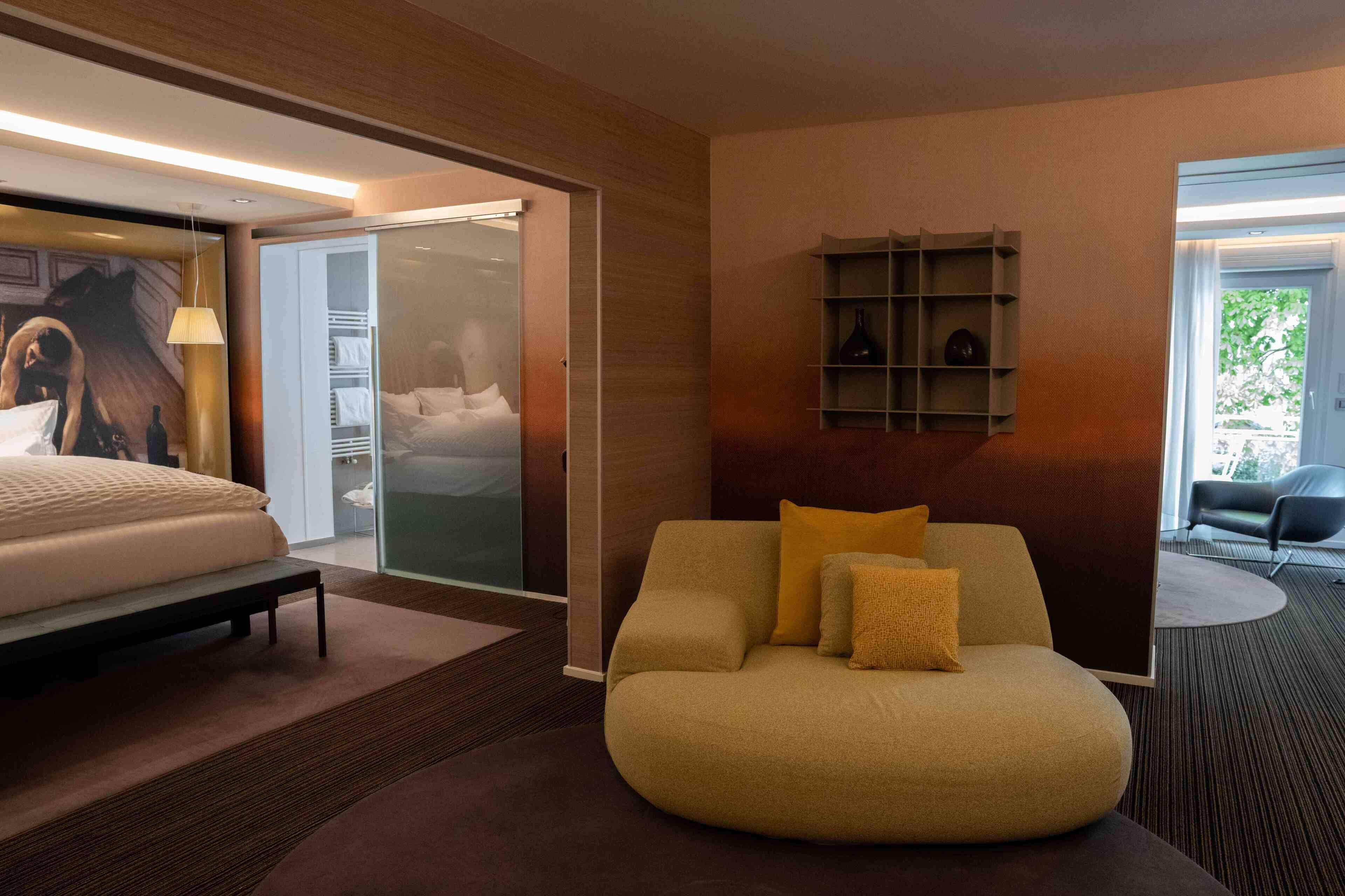 A modern hotel room with a bed, a sofa, a glass door leading to a balcony, and a shelf with decorative items.