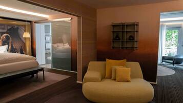 A modern hotel room with a bed, a sofa, a glass door leading to a balcony, and a shelf with decorative items.