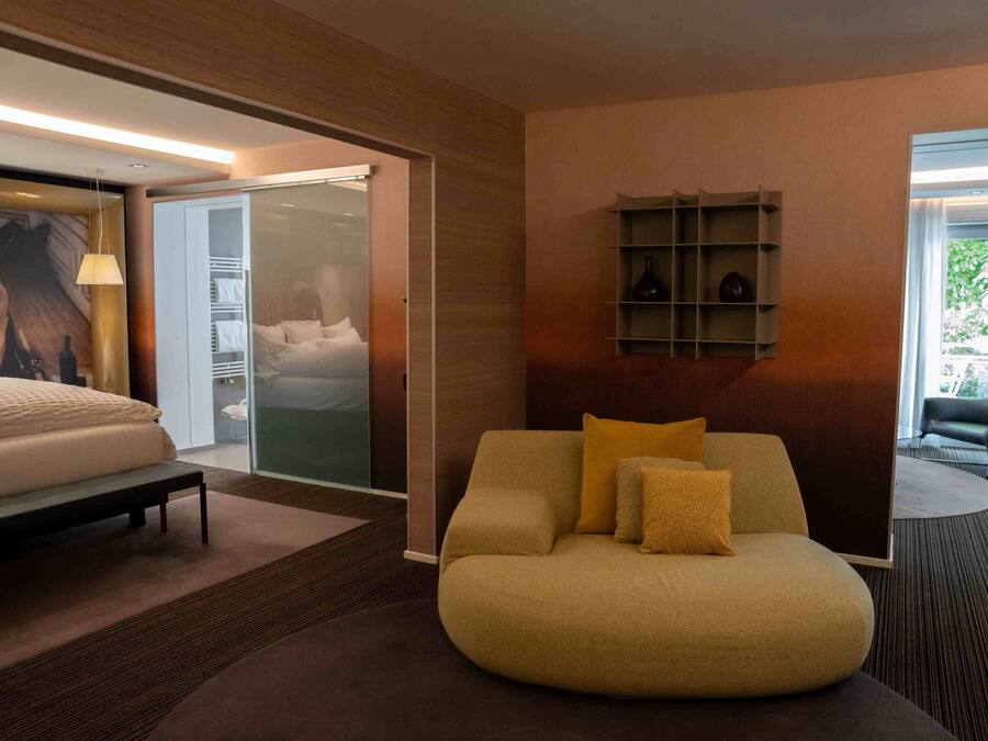 A modern hotel room with a bed, a sofa, a glass door leading to a balcony, and a shelf with decorative items.