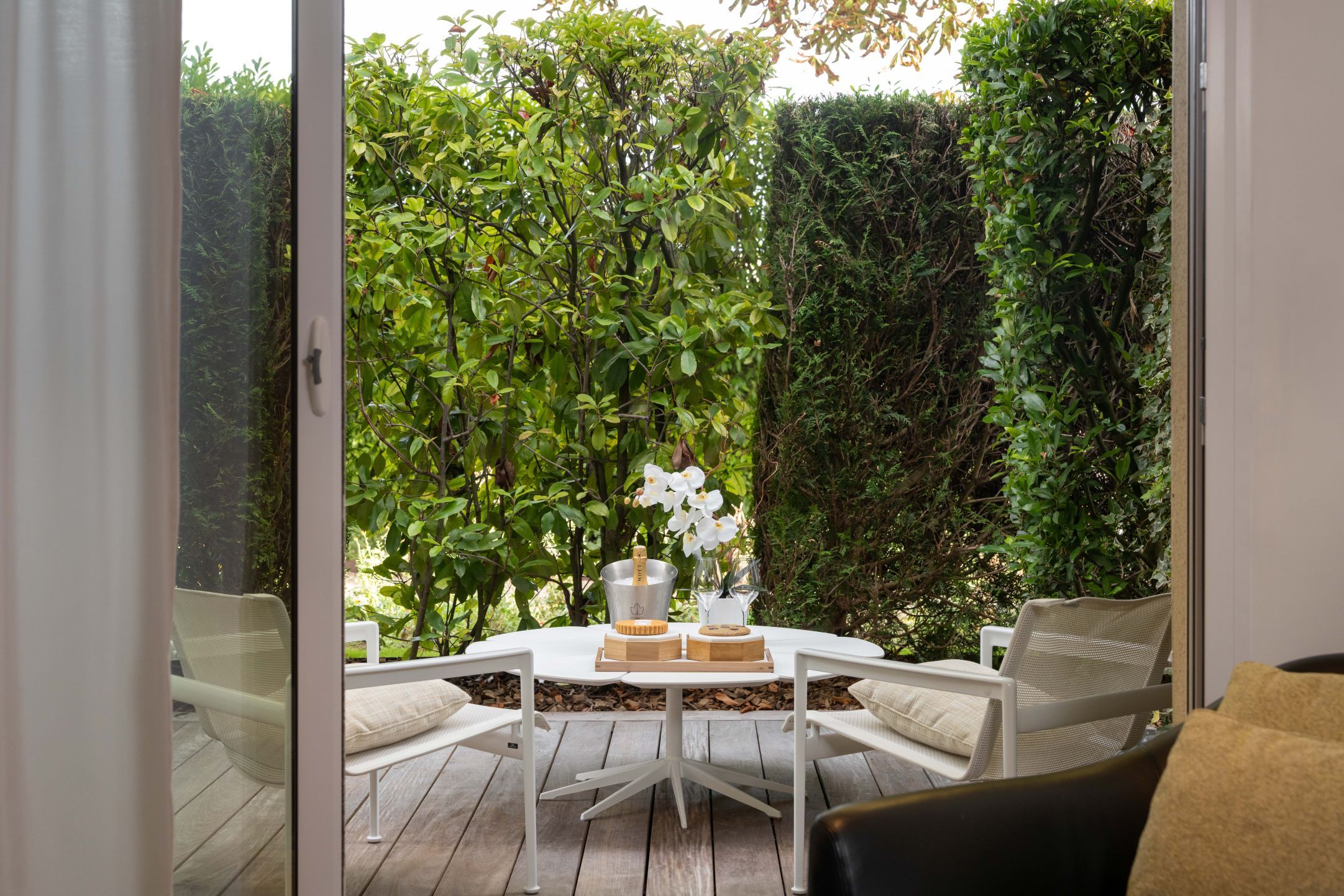 An outdoor patio with a round table, white chairs, and a bottle of champagne.