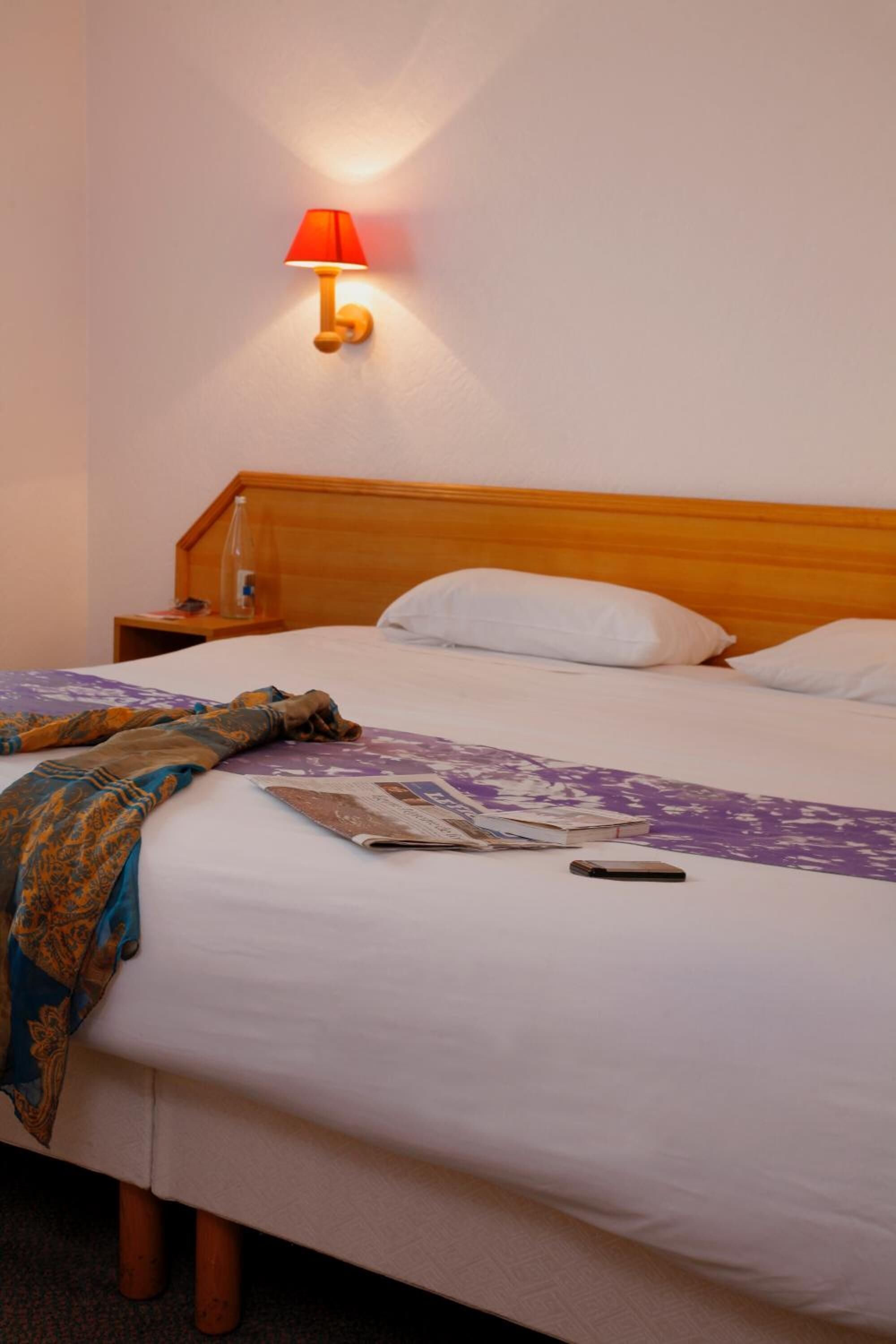 A hotel room with a wooden headboard, a bedside table with a bottle, a blanket, and a remote control on the bed.