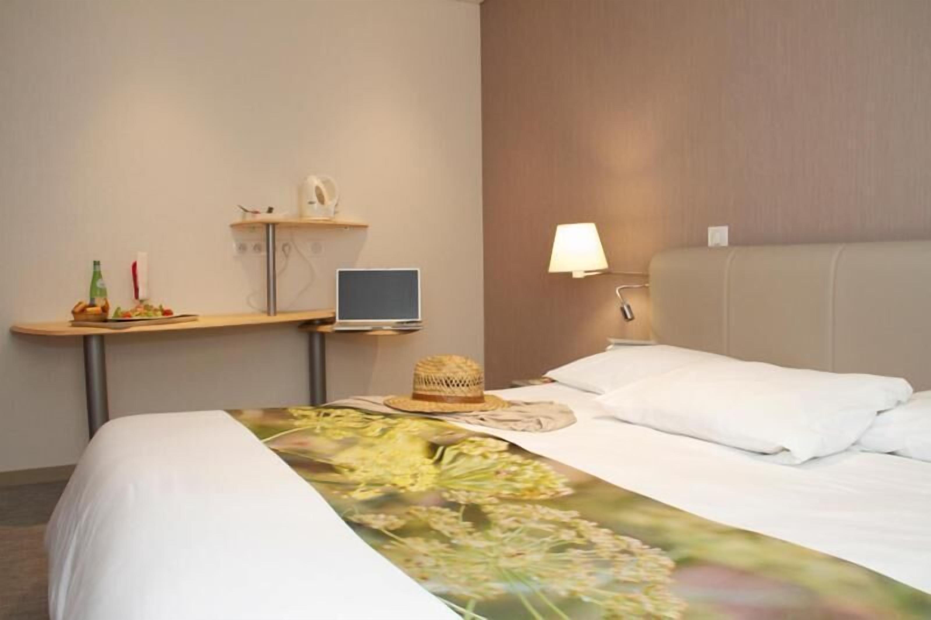A hotel room with a bed, a desk with a laptop, a lamp, and a small shelf with items.