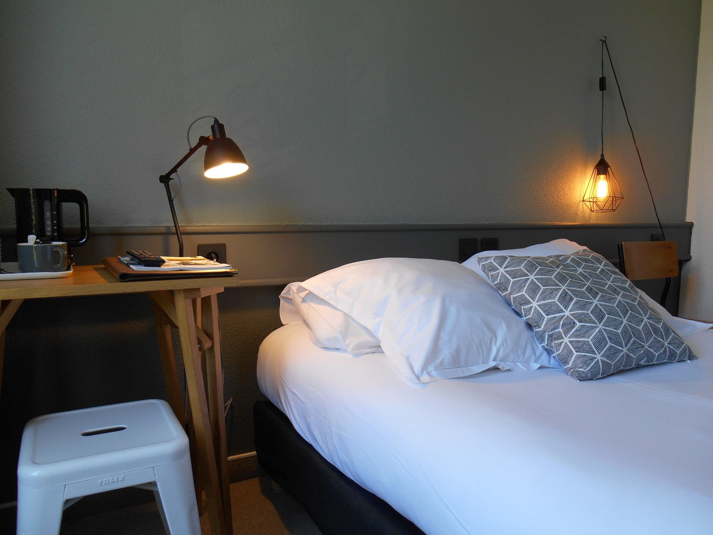 A neatly made bed with white and patterned pillows, a bedside table with a lamp, a coffee maker, and a small stool.