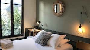 Ein ordentlich bezogenes Bett mit Kissen, ein Nachttisch mit Lampe, ein wandmontierter Spiegel und Blick auf Grün durch ein Fenster.