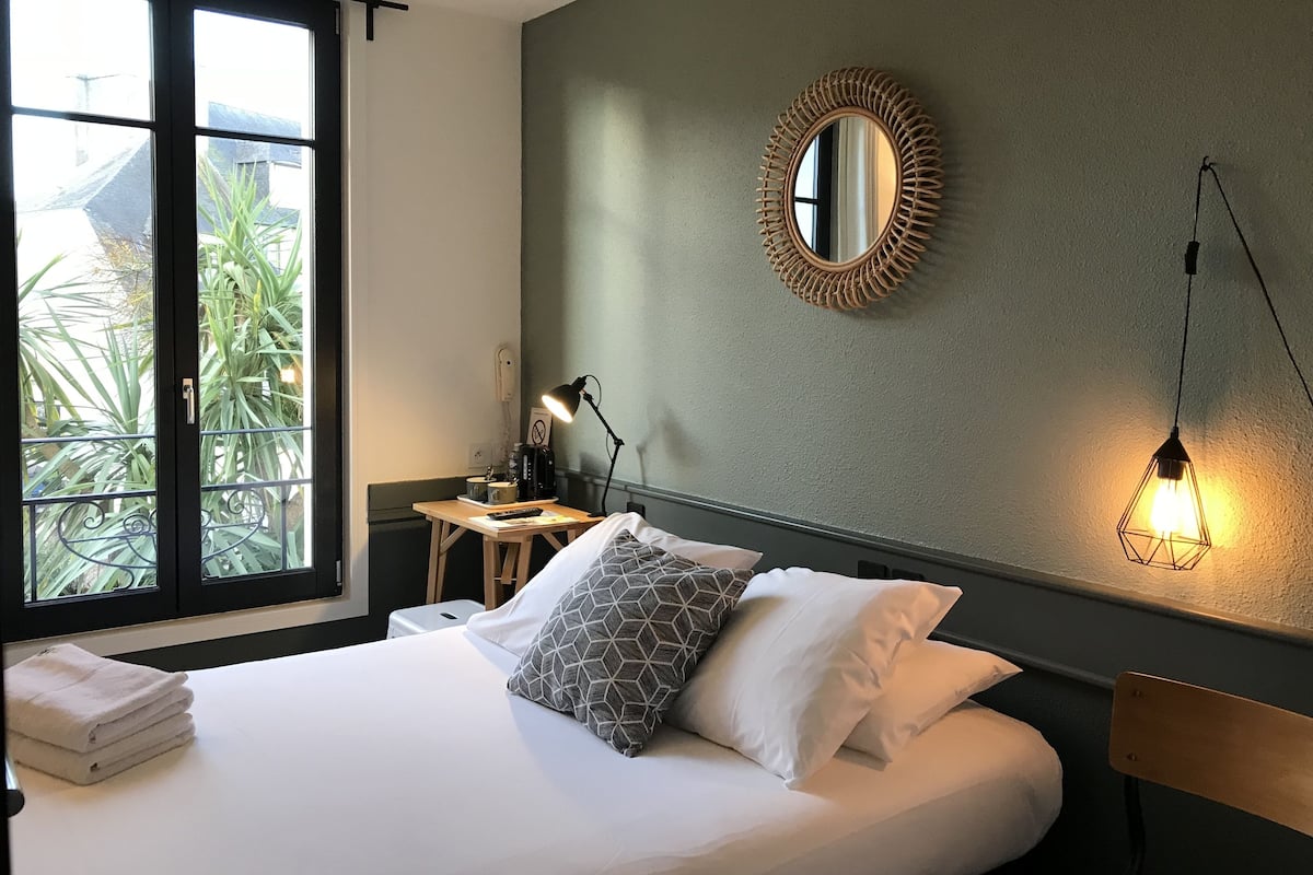 Een net opgemaakt bed met kussens, een nachtkastje met een lamp, een spiegel aan de muur en uitzicht op groen door een raam.