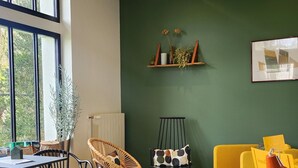 Ein modernes Wohnzimmer mit grüner Wand, einem gelben Sofa, einer grünen Couch und einem Essbereich mit Korbstühlen und einem Tisch.
