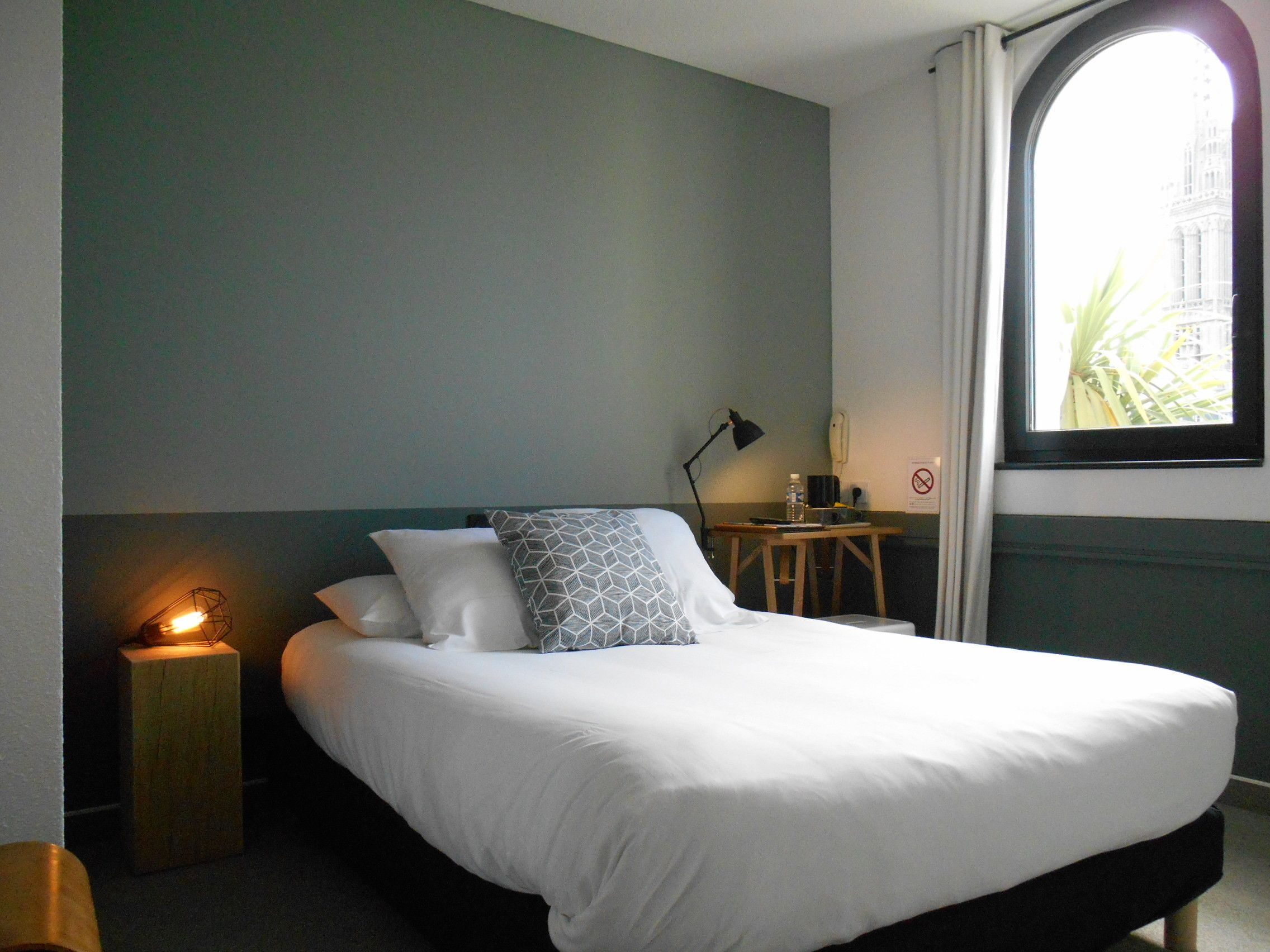 Una cama bien hecha con ropa blanca y un cojín estampado con motivos geométricos, una lámpara de noche, una mesita de madera con una lámpara, y vista a un edificio a través de una ventana.