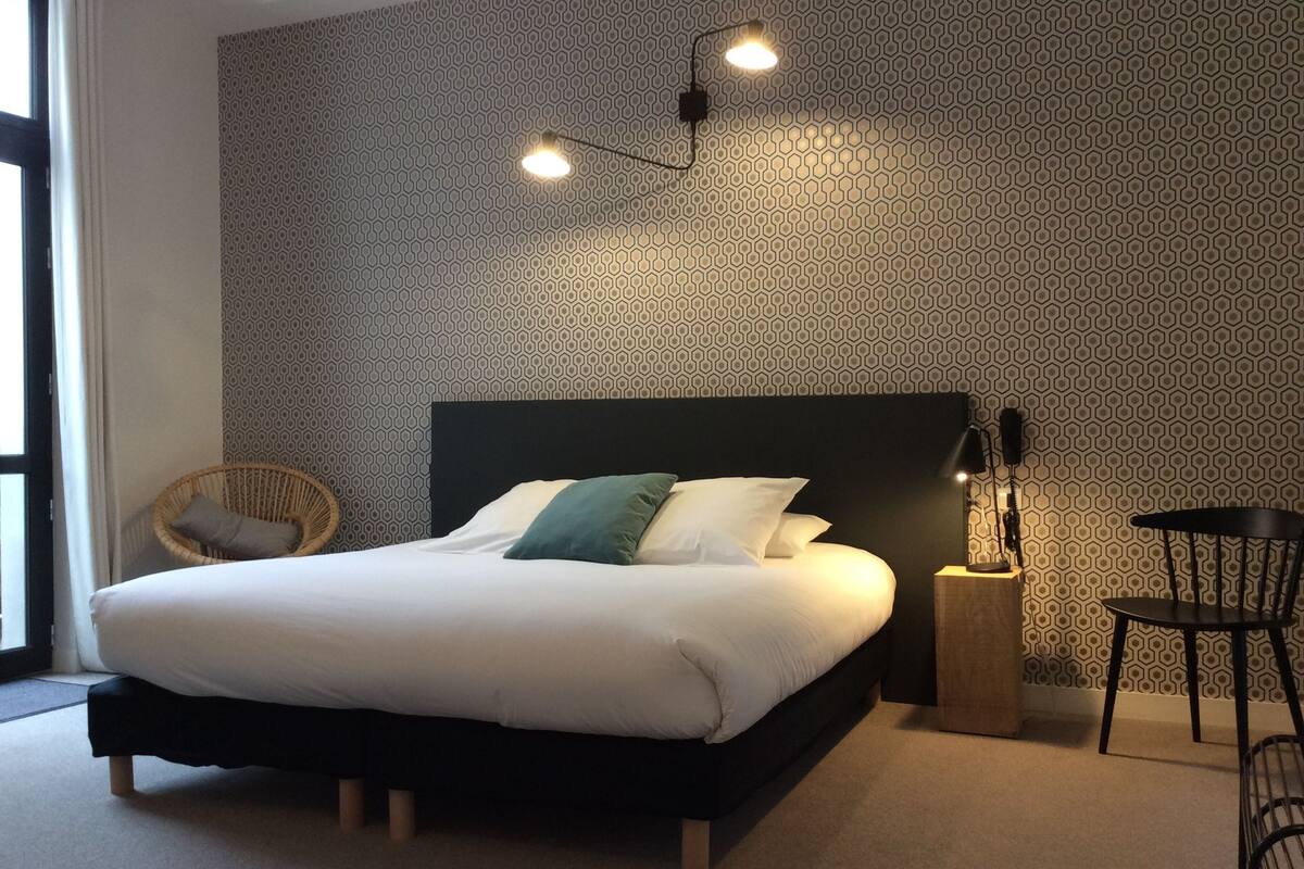 Een keurig opgemaakt bed met een zwart hoofdbord, een rieten stoel en een kleine tafel met een lamp.