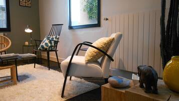 A living room with a white armchair, a wooden side table, a small wooden stool, a white fluffy rug, a lamp, and a framed date on the wall.