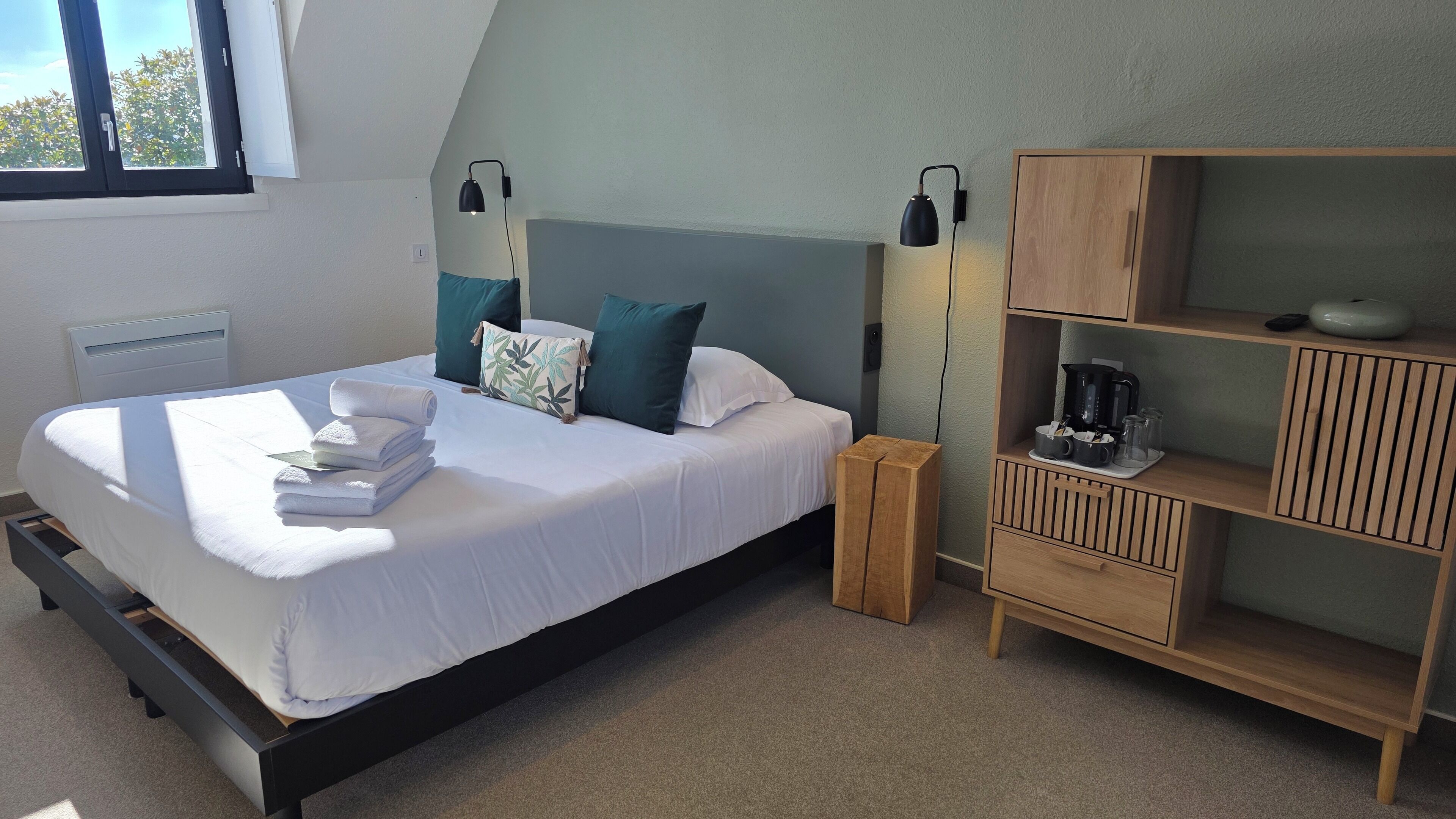 A neatly made bed with white linens and a stack of folded towels, a bedside table with a lamp, and a wooden nightstand.