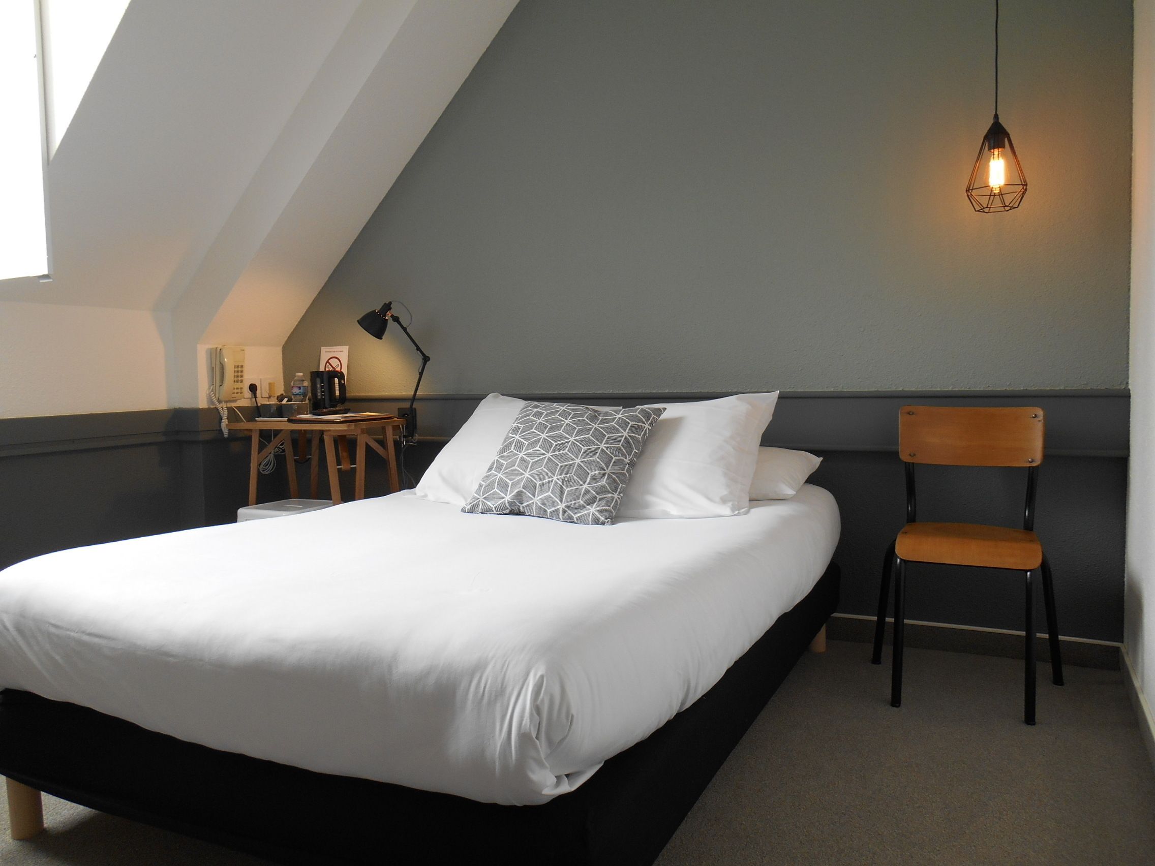 A neatly arranged bedroom with a bed, a chair, a desk with a lamp, and a hanging light fixture.