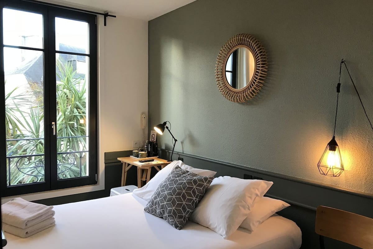 Een hotelkamer met een bed, een bureau, een stoel, een lamp, een spiegel en uitzicht op groen.