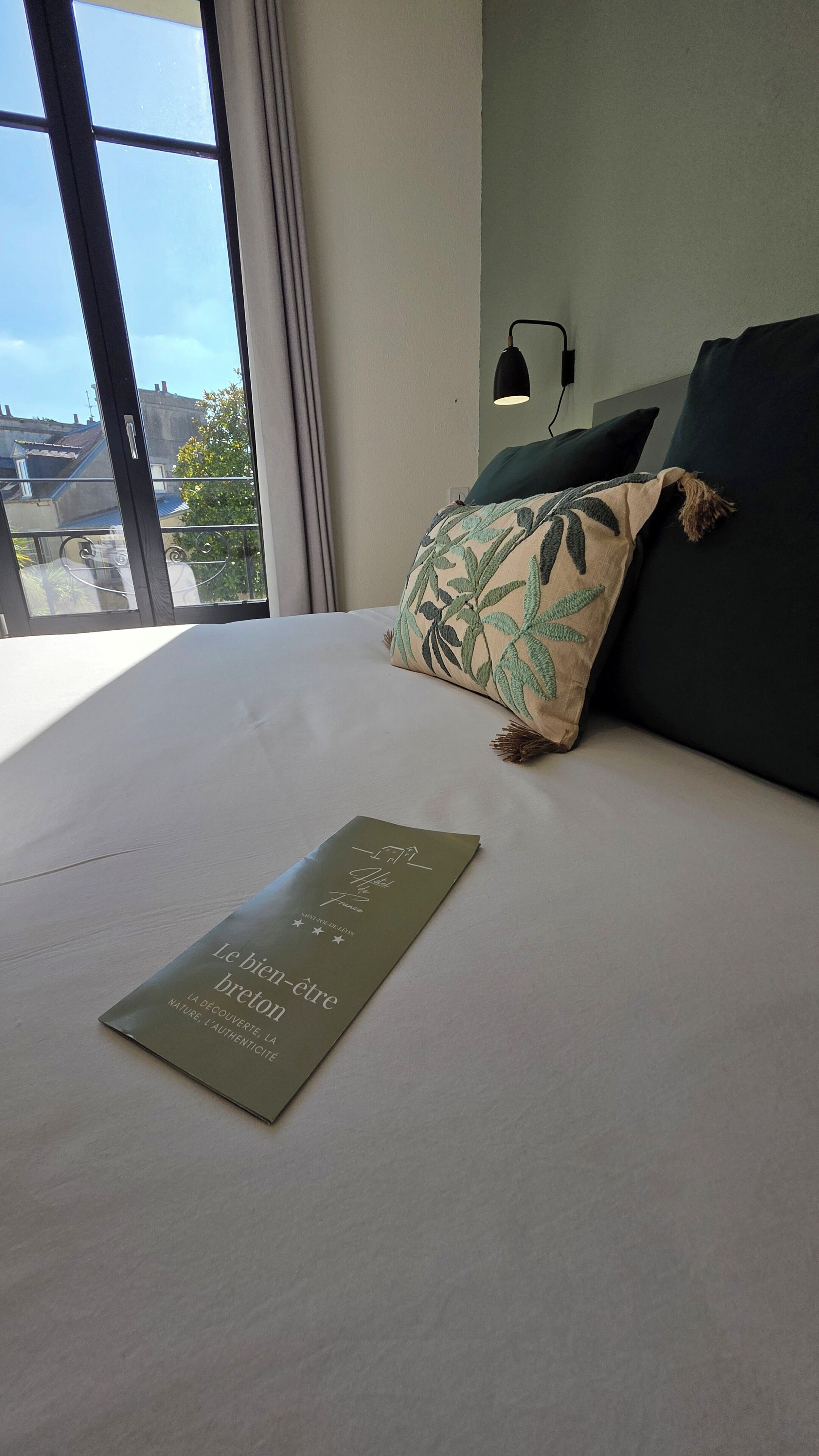 A neatly made bed with a green booklet on it, a dark green pillow, and a black lamp on the wall.