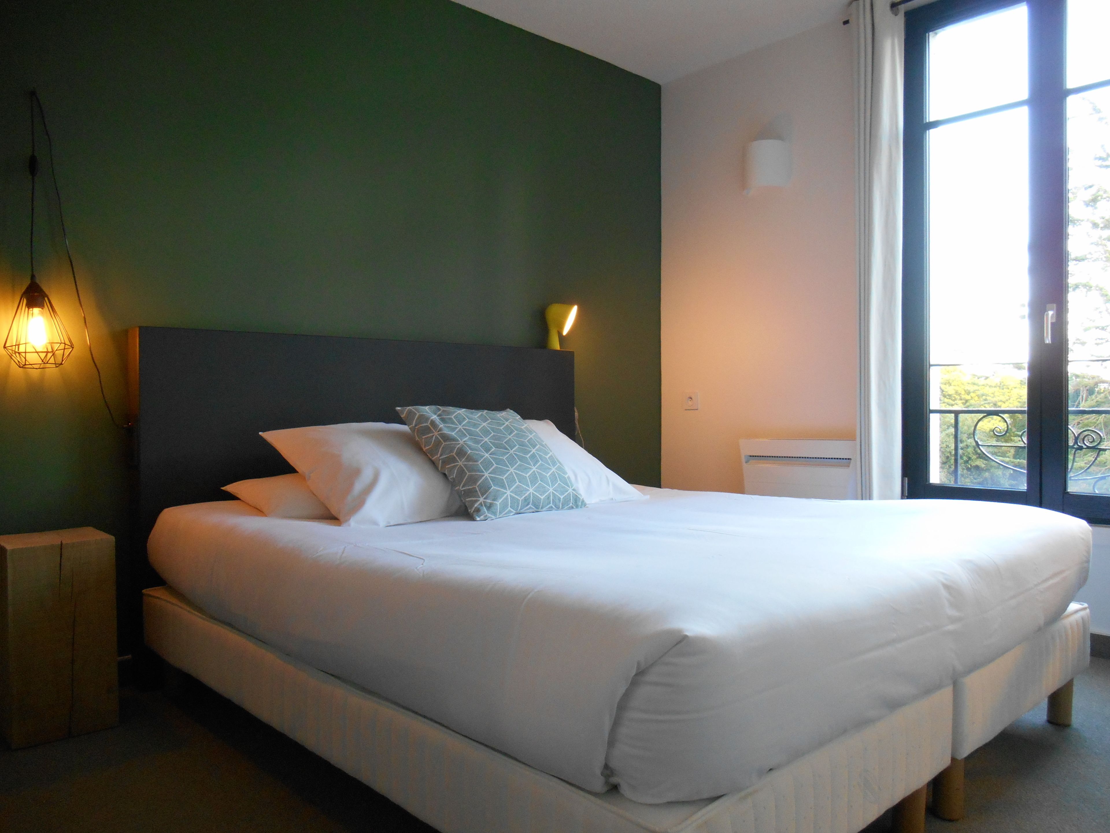 A neatly made bed with white linens and a geometric-patterned pillow, a bedside lamp, and a view of a balcony through a window.