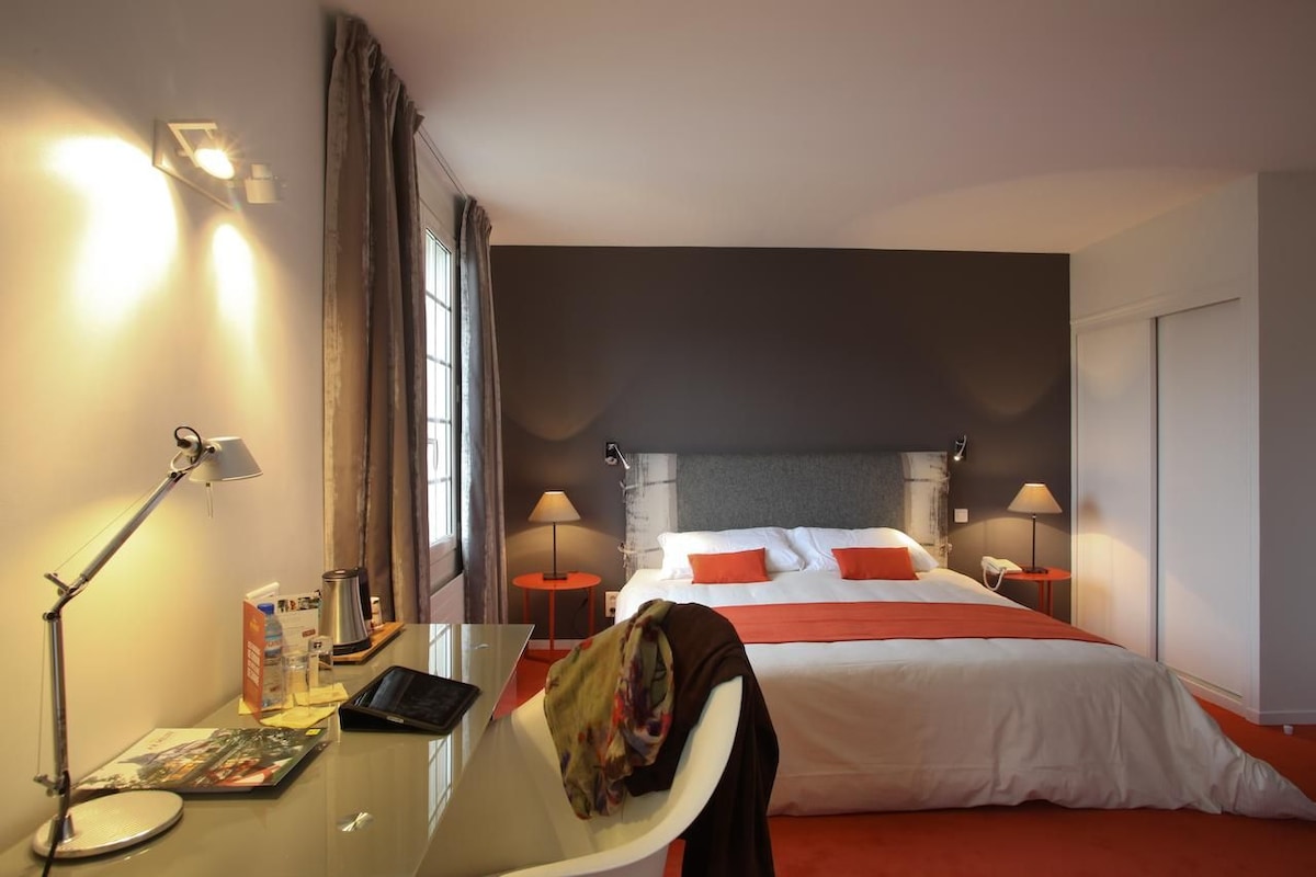 A hotel room with a bed, two bedside tables, a desk with a lamp, a chair, and a window with curtains.