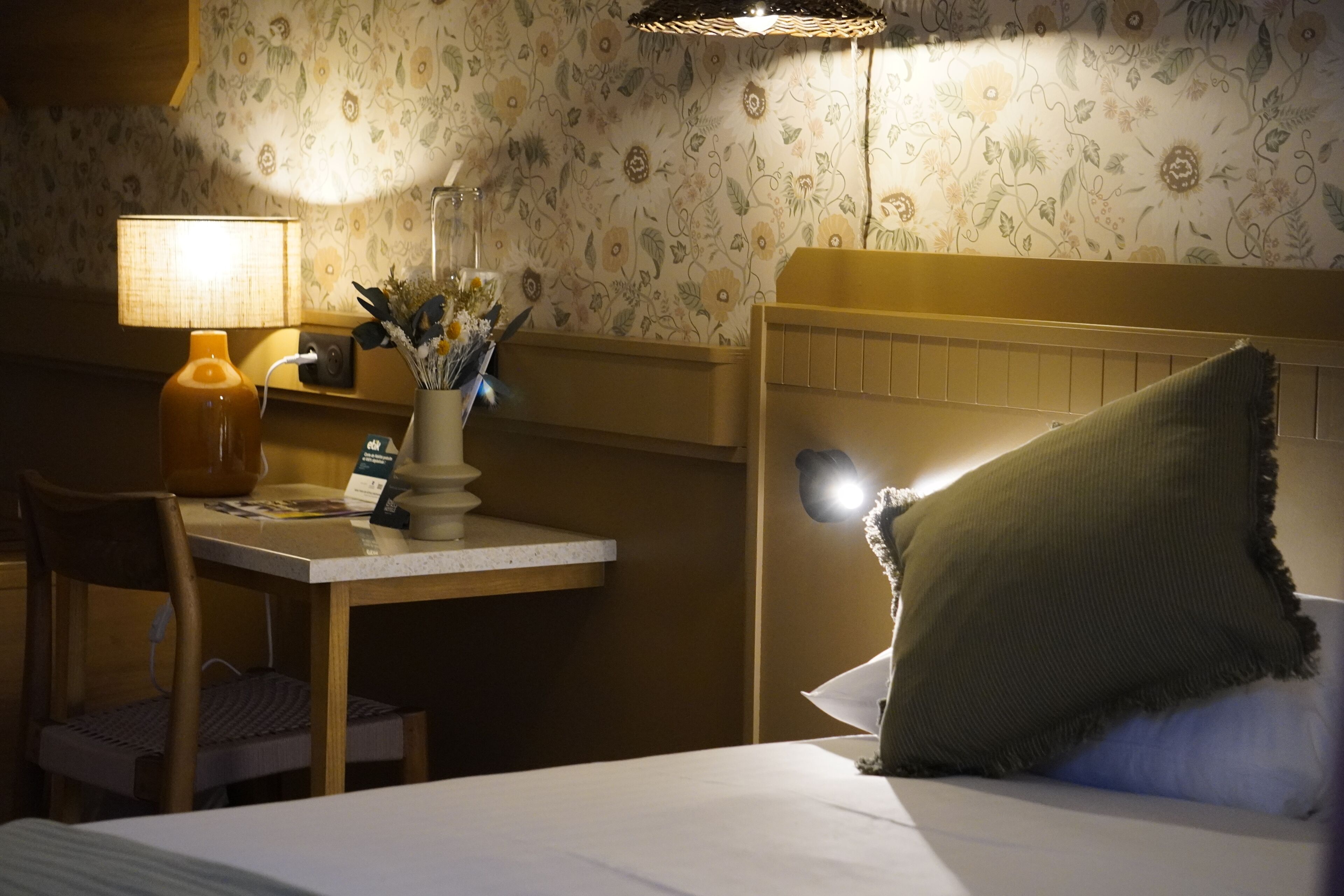A hotel room with a bed, a desk with a lamp, a chair, and a decorative vase with flowers.