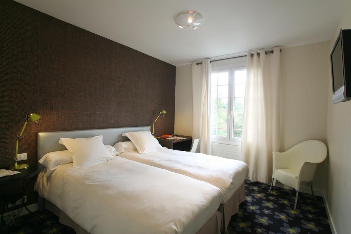 A hotel room with two beds, a desk with a lamp, a chair, and a window with curtains.