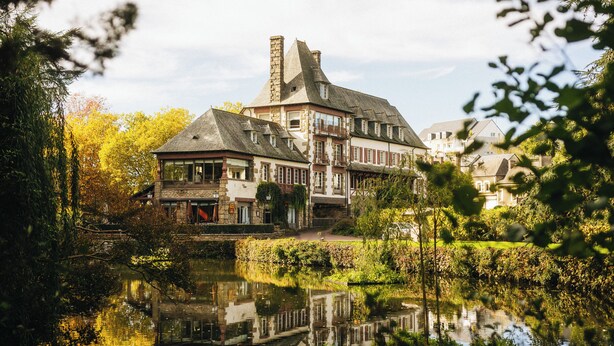 A traditional European-style building with a stone facade, multiple windows, and a prominent chimney. It is situated beside a calm body of water, surrounded by lush greenery and trees. - Hotel Ar Milin (Chateaubourg)