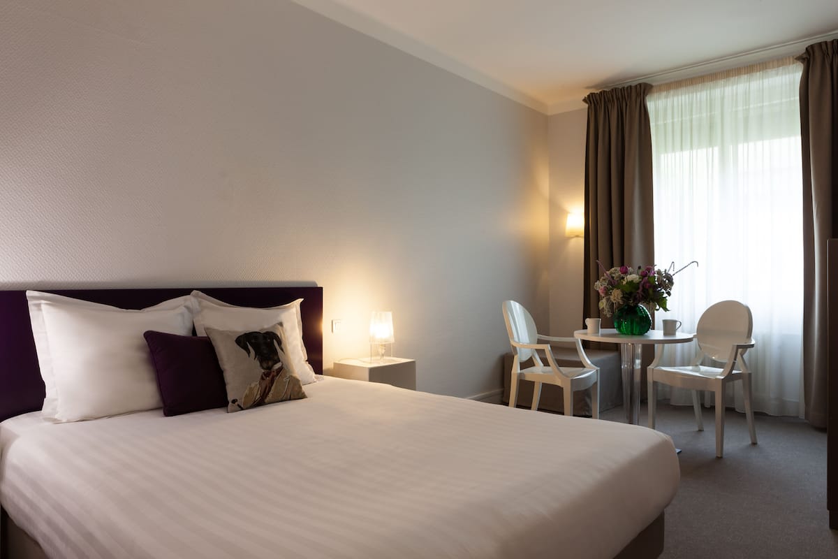 A hotel room with a large bed, a desk with a chair, a small table with a vase of flowers, and a window with curtains.
