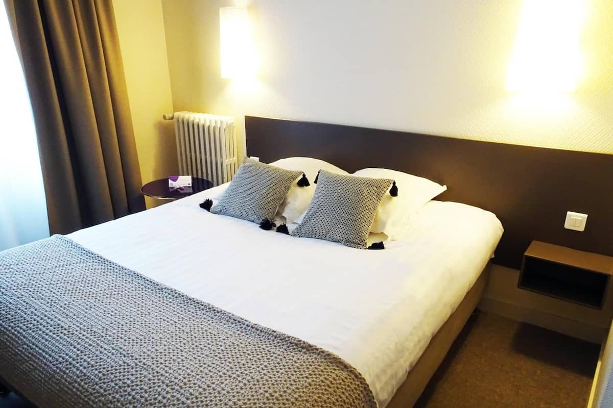 A hotel room with a large bed, two bedside tables, a nightstand, a radiator, and a wall-mounted light fixture.