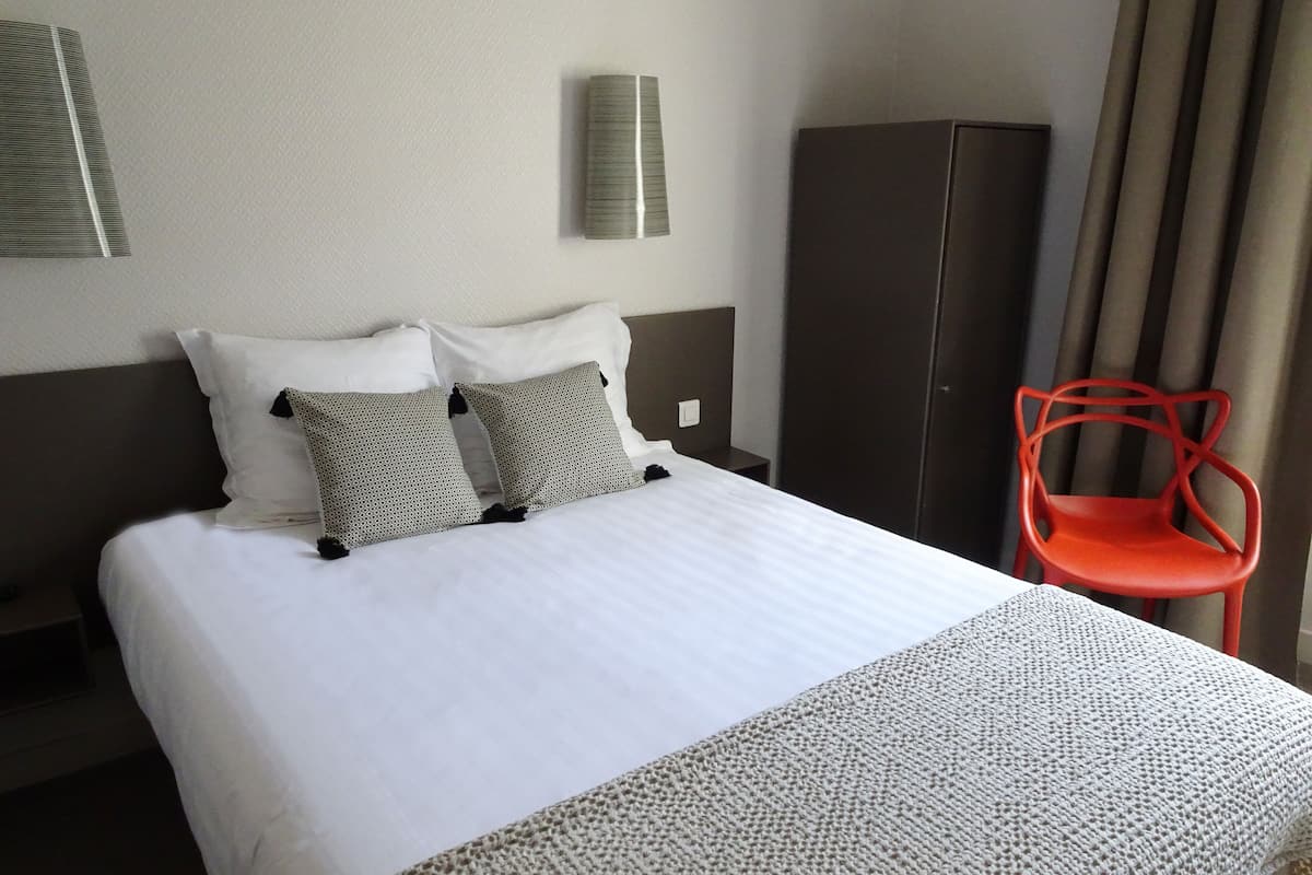 A hotel room with a large bed, two pillows, a red chair, a wardrobe, and a wall-mounted lamp.