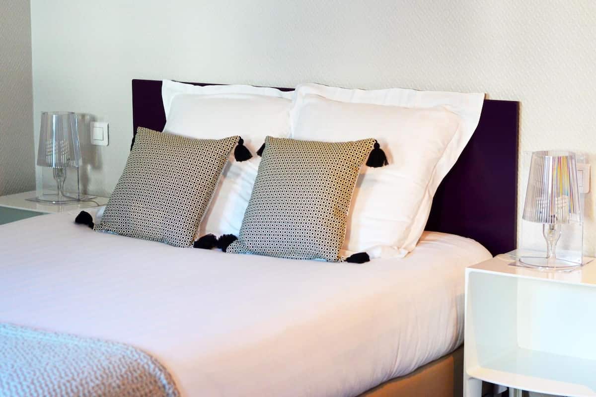 A neatly made bed with patterned pillows and a white duvet, a bedside table with a lamp, and two glasses.