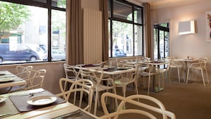 Une salle à manger d’hôtel avec de nombreuses tables et chaises, de grandes fenêtres avec des rideaux et une vue sur la rue.