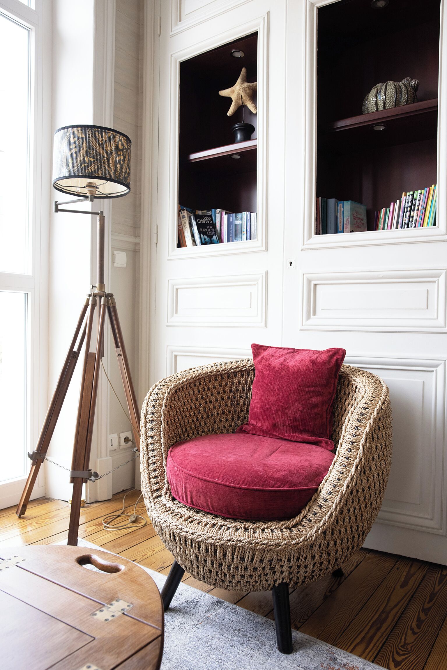 A wicker chair with a red cushion in a room with white paneled walls, a floor lamp, and shelves with books and decorative items.