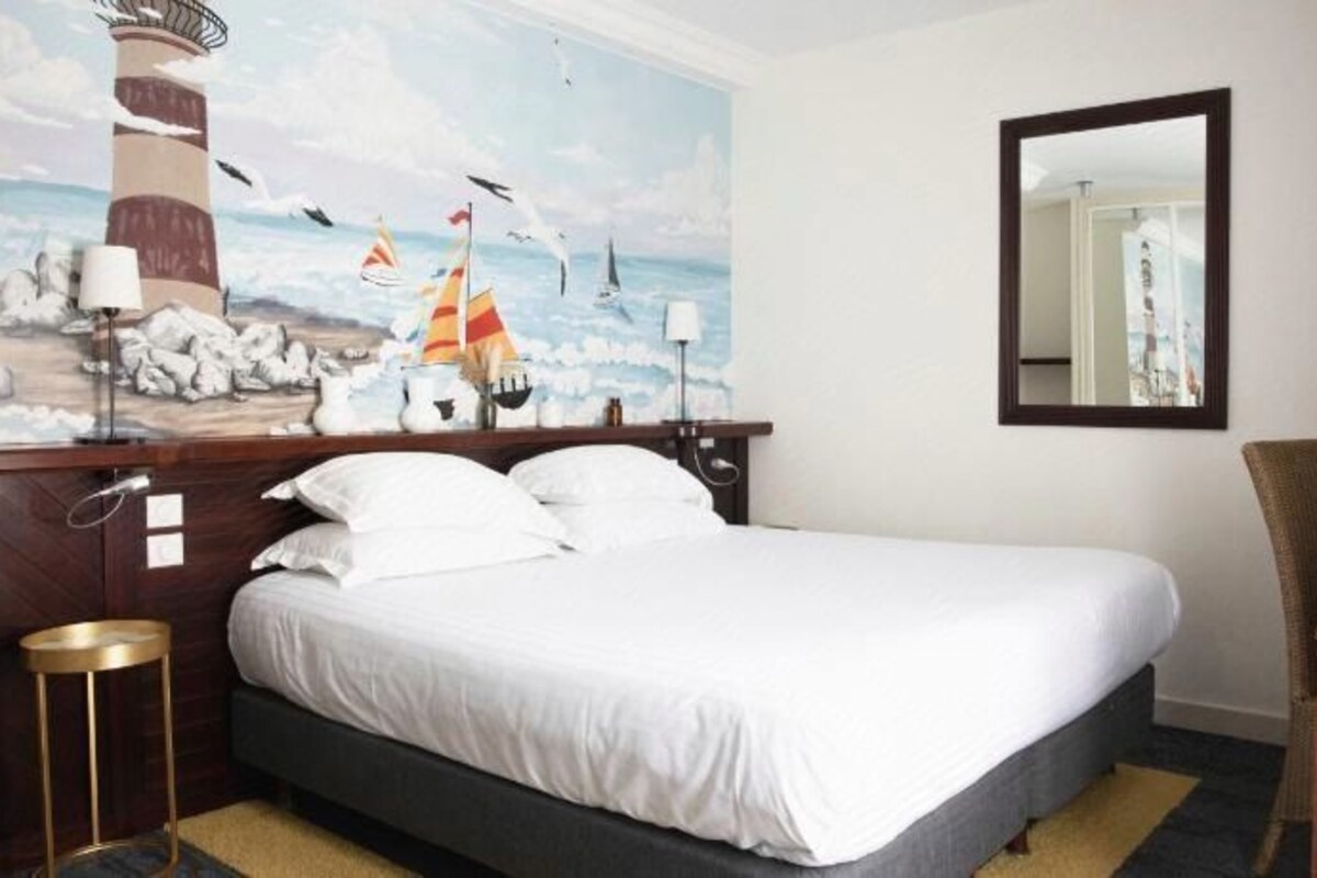 Une chambre d’hôtel avec un lit, un bureau, une chaise, un miroir et un tableau représentant un phare et des voiliers accroché au mur.