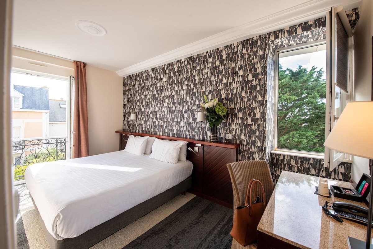 Een hotelkamer met een bed, een bureau met lamp, een stoel, een raam met uitzicht en behang met patroon.