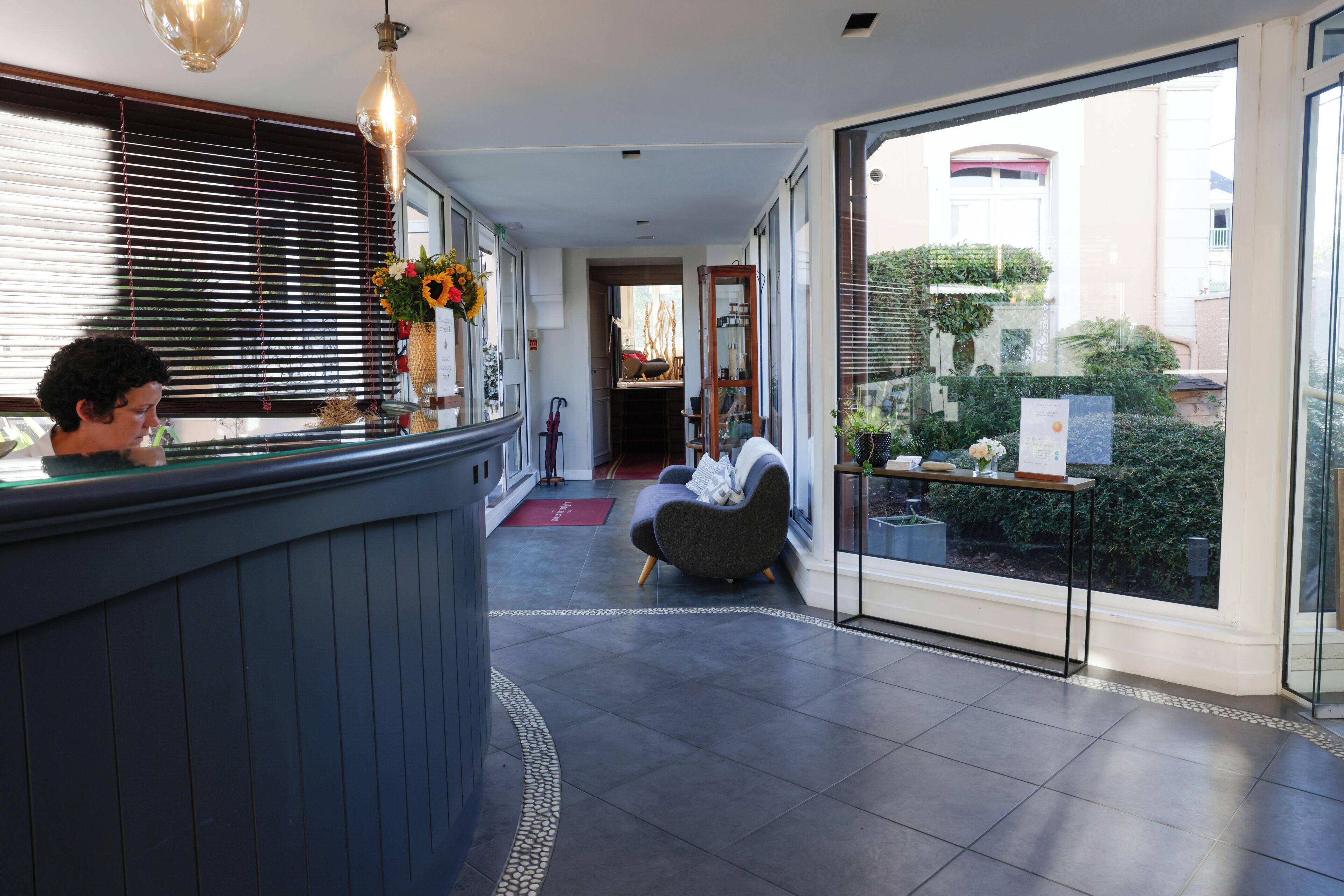 A modern hotel lobby with a reception desk, seating area, and large windows offering views of the surrounding buildings and greenery.