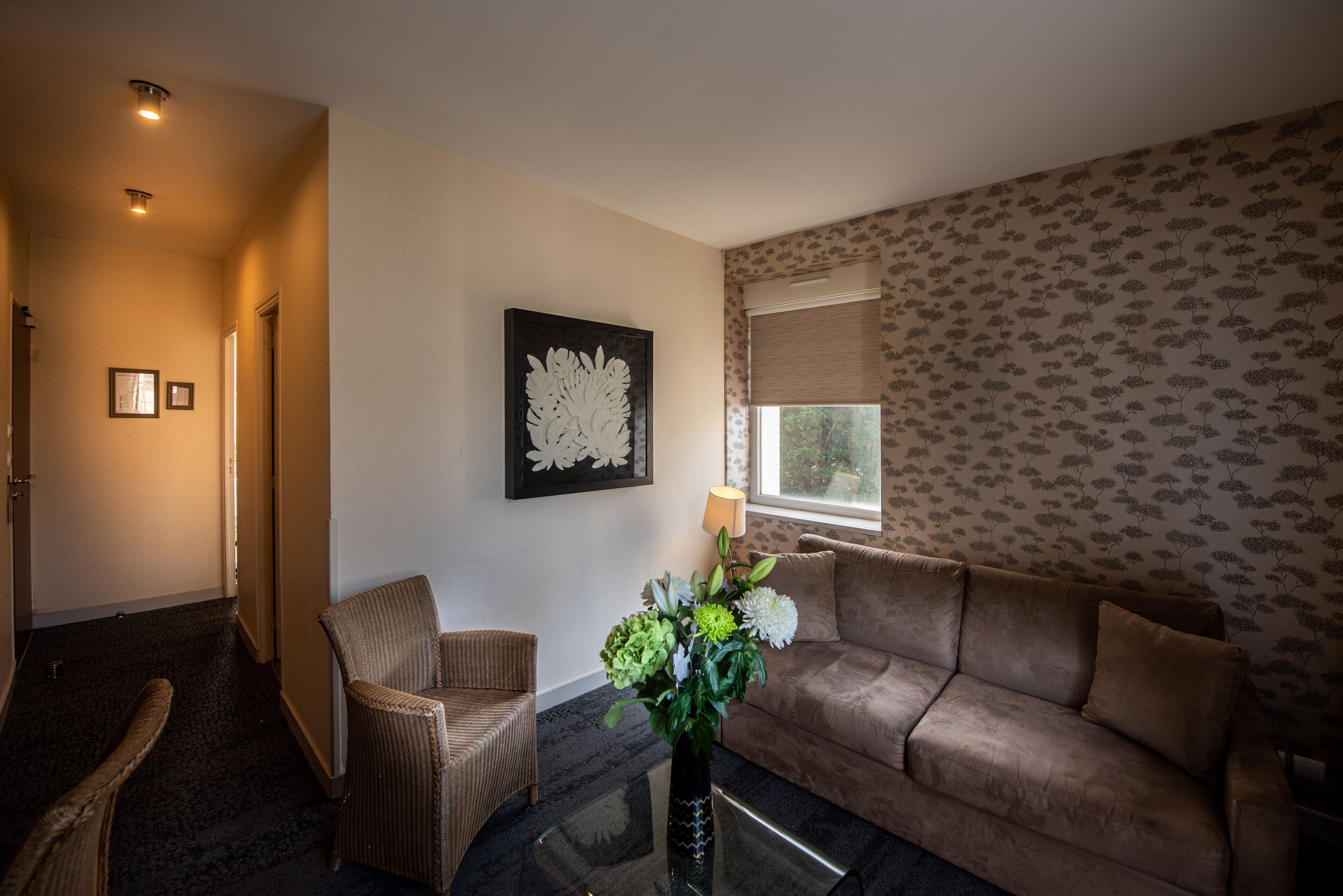 Una sala de estar con un sofá marrón, una silla de mimbre, una mesa de centro de vidrio y un jarrón con flores. En la pared hay una obra de arte enmarcada y una ventana con cortina estampada.