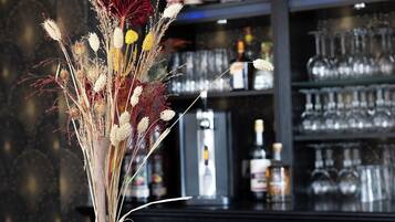 Zona de bar con un jarrón de flores secas en la barra, una variedad de botellas y vasos en estanterías, y lámparas colgantes.