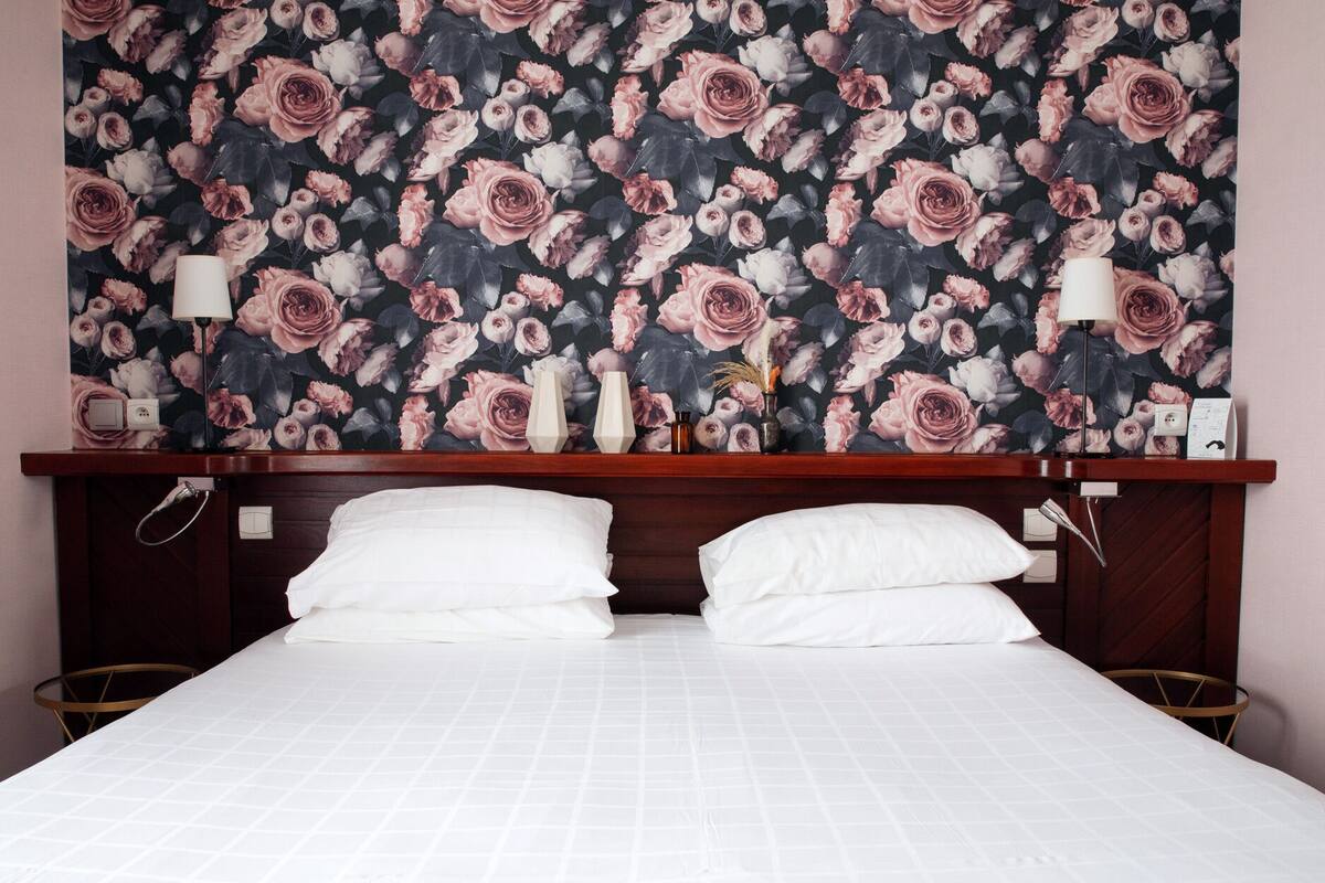 Een hotelkamer met een groot bed, twee witte kussens, een houten hoofdbord en behang met bloemen.