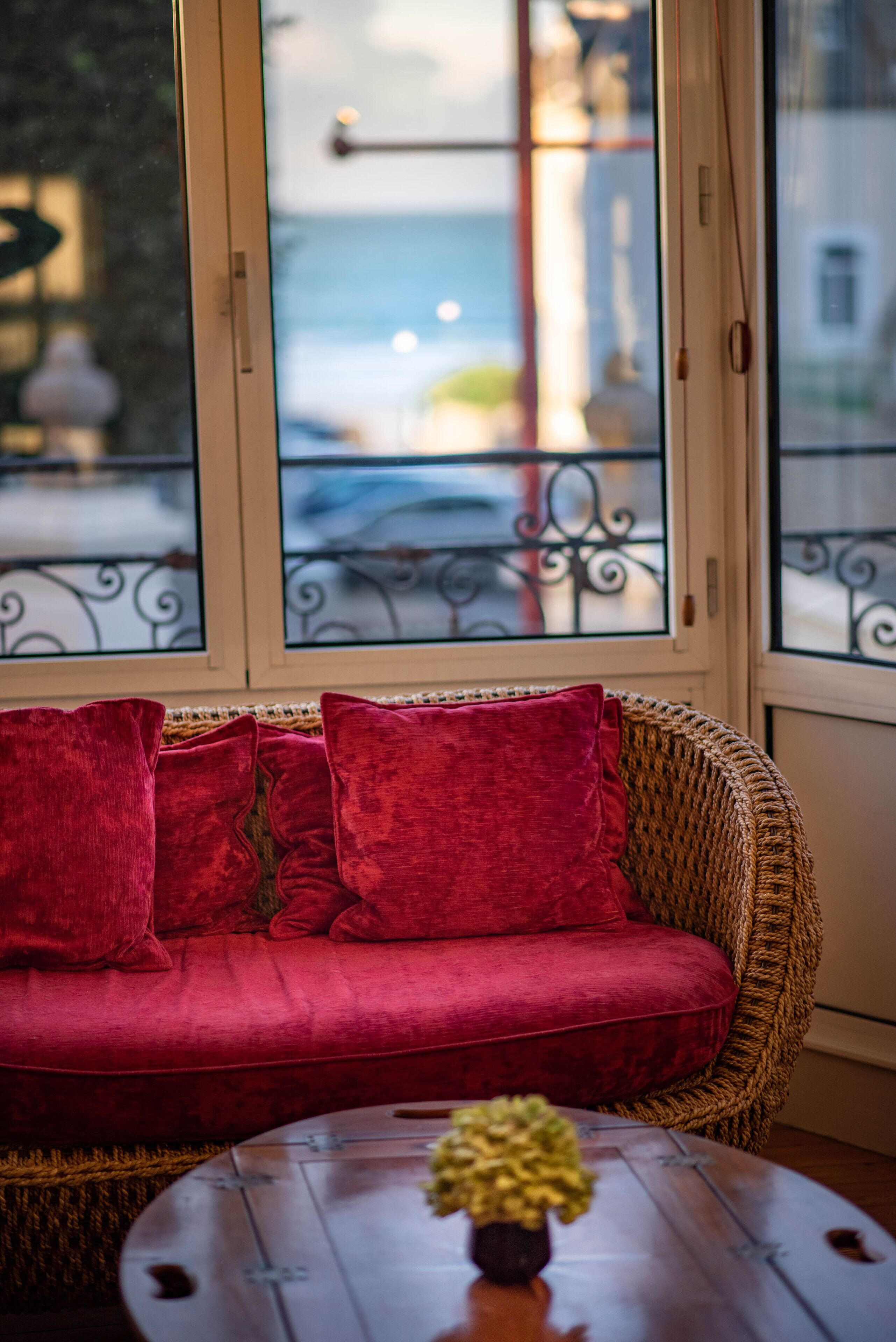 A wicker sofa with red cushions, a round wooden table with a vase, and a view of a balcony with metal railings.