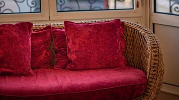 A wicker sofa with red cushions, a round wooden table with a vase, and a view of a balcony with metal railings.