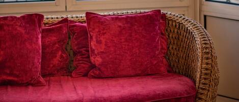 Un sofá de mimbre con cojines rojos, una mesa redonda de madera con un florero, y vistas a un balcón con barandillas de metal.