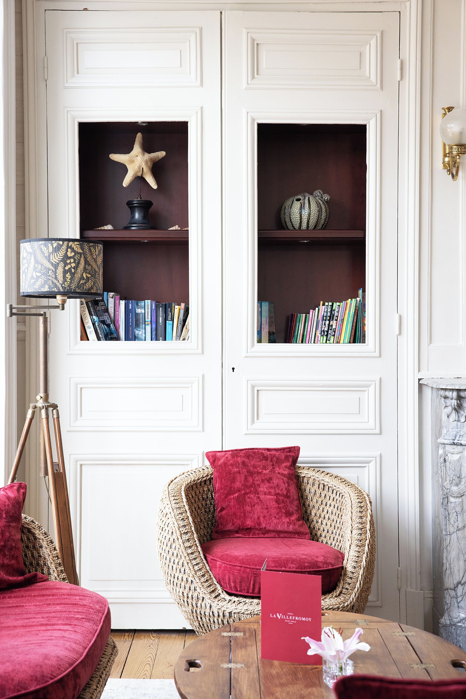 Una stanza con una poltrona in vimini, un cuscino rosso, una libreria piena di libri, una decorazione a stella marina e una lampada.