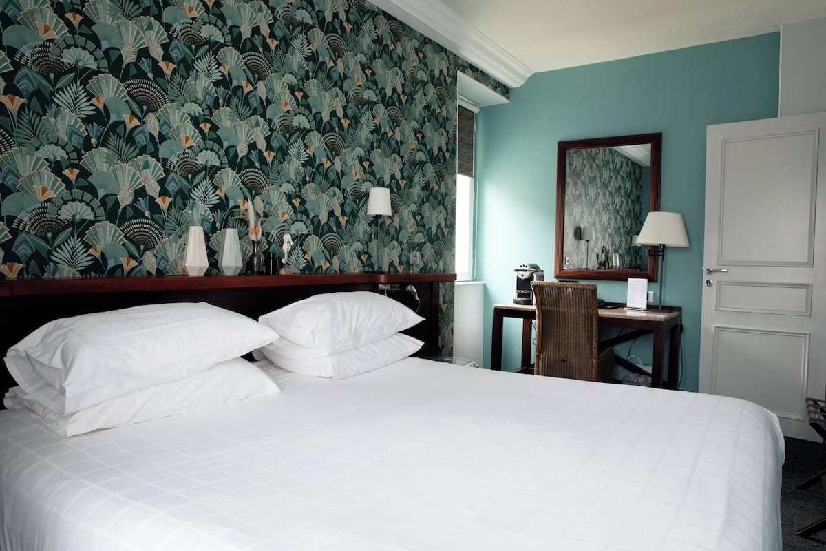 Een hotelkamer met een groot bed, een bureau met stoel, een spiegel en een muur met een bladeren patroon.