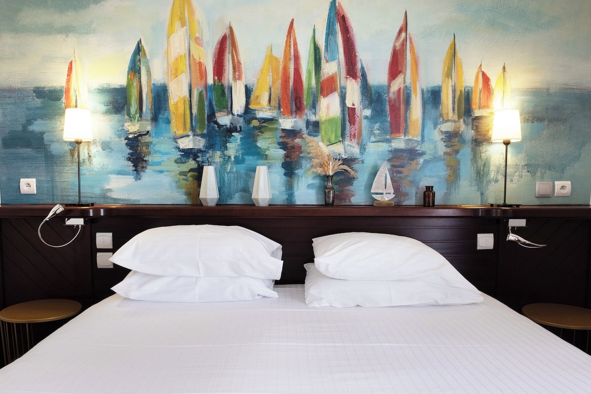 Een hotelkamer met een groot schilderij van kleurrijke zeilboten aan de muur, twee bedlampen, een bed met witte lakens en een klein rond tafeltje.