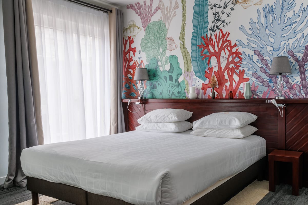 Een net opgemaakt bed met witte lakens in een hotelkamer, met een houten hoofdbord, een wanddecoratie van koraal en zeeleven, en een klein nachtkastje met een lamp.